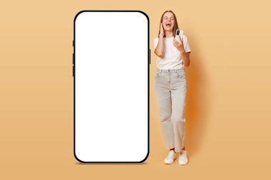Gündelik kıyafetler giyen genç beyaz kadın akıllı telefon reklamları okuyarak büyük bir cep telefonunun yanında boş boş duruyor.