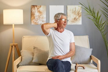 Yaşlı bir adamın boynu ağrıyor. Son sınıflarda kas krampı. Yaşlılıkta omurga sorunları. Yorgun ve ağrılı kıdemli. Oturma odasındaki kanepede oturan, boynu ağrıyan üzgün yaşlı adam.