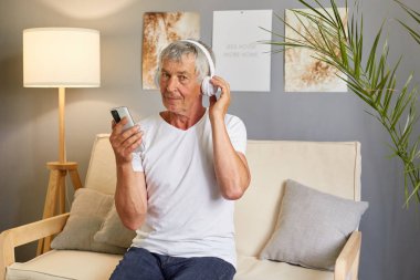 Öksürük üzerinde beyaz tişört giyen, elinde cep telefonu ve kulaklığıyla oturmuş müzik ya da sesli kitap dinleyen ve evde boş vakitlerin tadını çıkaran yaşlı ve düşünceli bir beyefendi..