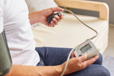 Beyaz tişört ve kot pantolon giyen, modern tonometre kullanan, kan basıncını ölçen, oturma odasındaki kanepede otururken kendini kontrol eden tanınmamış bir erkek hasta..