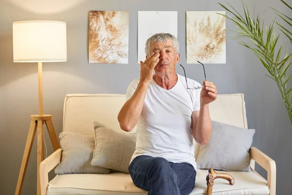 Tired senior with glasses. Sore eyes from infection. mature old man wearing white T-shirt rubbing and wiping eyes sitting on sofa in living room home interior