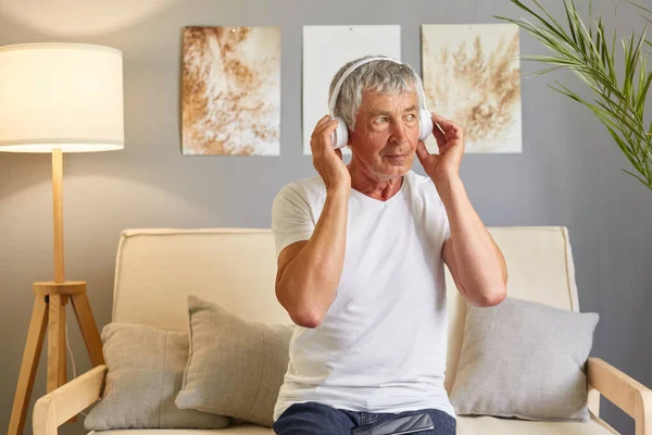 Older gentleman dressed in a white T-shirt using his mobile phone to listen to music online using modern headphones sitting on a sofa enjoying his free time listening to audio book.