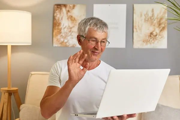 Happy old retired man waving hand talking to webcam make distant video call chat on laptop whiloe sitting on sofa, cheerful grandpa enjoy online communication technology looking at computer screen at home