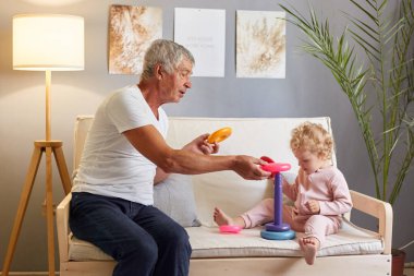 Büyükbaba ve torun. Eğlenceli bir bebek oyunu. Mutlu aile hayatı. Tatlı kız oynuyor. Beyaz saçlı adam, bebek bebekle oyuncak oynuyor oturma odasında kanepede.