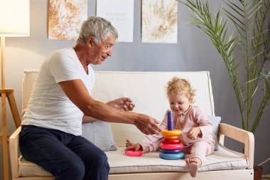 Beyaz t-shirt giyen, mutlu, gri saçlı, olgun bir adam. Oturma odasında küçük torunuyla kanepeye oturmuş, çok renkli piramitlerle oynuyor..