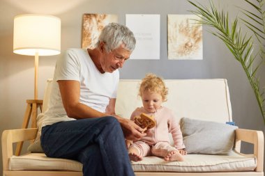 Beyaz t-shirt giyen hoş bir adam, bebeğiyle birlikte oturma odasında koltukta oturuyor. Küçük tatlı torununa yeni tahta oyuncaklar getiriyor..