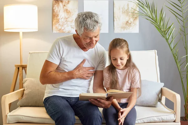 Little girl and her grandfather smiling readinjg interesting book enjoying fairy tale sitting on couch in living room at home interior wearing casual clothing.