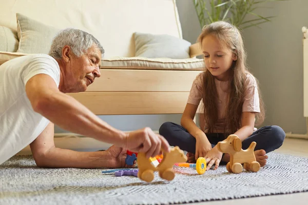 Mature man wearing white t-shirt playing with wooden toys with his granddaughter posing in living room sitting on floor family enjoying free time together.