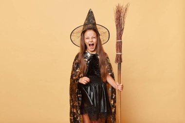 Cadı kostümü giymiş çılgın bir kız bej arka planda izole edilmiş süpürge tutuyor cadılar bayramı kutlamasında eğleniyor ve ağzı sonuna kadar açık bir şekilde bağırıyor..