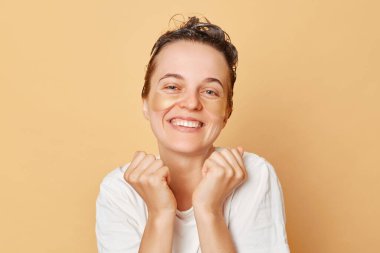 Neşeli pozitif kadın, beyaz tişörtlü, gözünün altında hidrojel yamalar ve kafasında şampuan köpüğü olan bej arka planda izole edilmiş güzel bir sabah rutininin keyfini çıkarıyor..