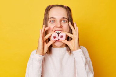 Atlı atkuyruklu neşeli, neşeli genç kız sarı arka planda izole edilmiş şekerlemeleri ısırıyor kameraya bakıyor tatlıları yiyor..