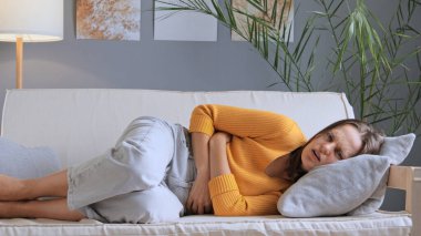 Mide ağrısından muzdarip, günlük kıyafetleri içinde mutsuz genç bir kadın, evdeki kanepede uzanmış, karnını tutuyor, karın ağrısı ve rahatsızlık çekiyor, regl oluyor..