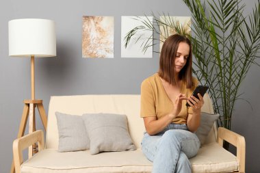 Akıllı telefon bağımlılığı. Cep telefonuyla serbest çalışma. İnternet üzerinden sosyal medya ağı. Blogcu çevrimiçi istatistikleri kontrol ediyor. Ciddi kahverengi saçlı, bej tişört giyen ev içinde telefon kullanan bir kadın.