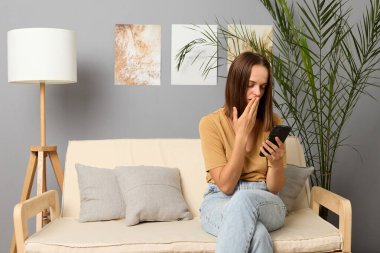 Şok içeriği. Cep telefonuyla arkadaşlarla sohbet etmek. Akıllı telefondan internet taraması. İnternetten alışveriş bağımlılığı. Şaşırdım. Kahverengi saçlı, bej tişörtlü kadın evde telefon kullanıyor..