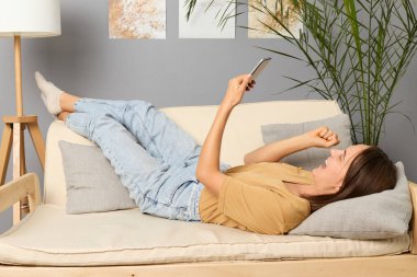 Dijital teknoloji. Dijital bir eğlence. Son derece mutlu, kahverengi saçlı, bej tişörtlü, kanepeye uzanmış, elinde akıllı telefon, yumrukları sıkılmış, oturma odasında neşeyle bahis oynayan ve kazanan bir kadın..