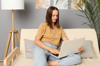 Laptop araştırması. Online iş. Ağ yöneticisi. Şirket çalışanı. Sakin, kahverengi saçlı, bej tişört ve kot giyen bir kadın koltukta oturuyor ve evdeki taşınabilir bilgisayarda çalışıyor..