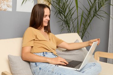 Çalışan şebekesi. Online tefekkür. Girişimci başarısı. Çekici, kahverengi saçlı, bej tişörtlü ve kot pantolonlu bir kadın kanepede oturuyor ve oturma odasında dizüstü bilgisayarla çalışıyor..
