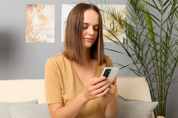 Web browsing on smartphone. Checking social media profiles online. Freelance work via smartphone apps. Confident brown haired woman wearing beige T-shirt using phone in home interior.