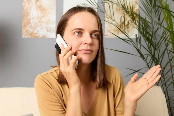 细胞连接 智能手机的电话 给朋友打电话通信工具 电话讨论 困惑的黑发女人穿着米色的T恤 坐在沙发上通过电话交谈 — 图库照片