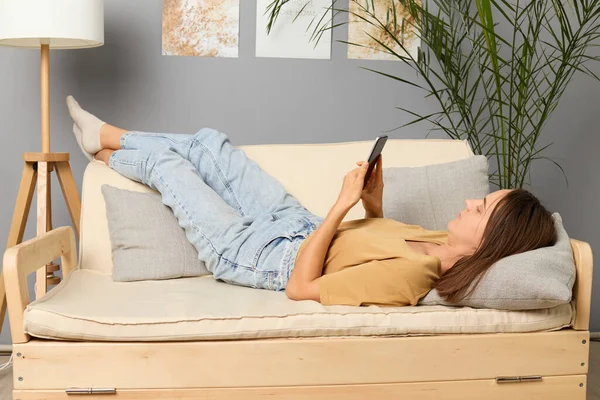 Digital technology. Online communication. Mobile app usage. Social network. Calm brown haired woman wearing beige T-shirt lying on sofa holding smartphone in living room.
