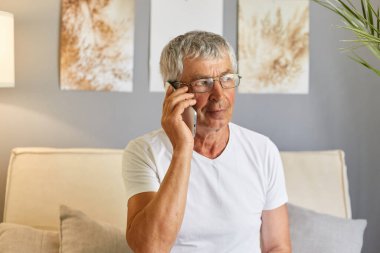 Sakin, gri saçlı, normal beyaz tişört giyen ve gözlüklü bir adam. Evdeki koltukta oturmuş akıllı telefon tutuyor ve arkadaşıyla cep telefonuyla konuşuyor..