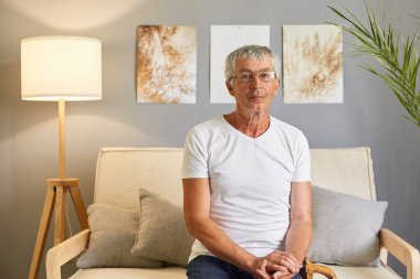 Ciddi, kendine güvenen, beyaz tişört giyen ve gözlüklü olgun bir adam. Evdeki koltukta oturmuş, kanepede oturmuş konsantre bir ifadeyle kameraya bakıyor..