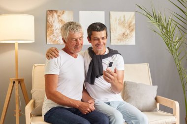 Mutlu gri saçlı baba ve yetişkin oğul oturma odasındaki kanepede oturup selfie çekerken akıllı telefon kullanıyor ve internet iletişimi için cep telefonu kullanıyor..