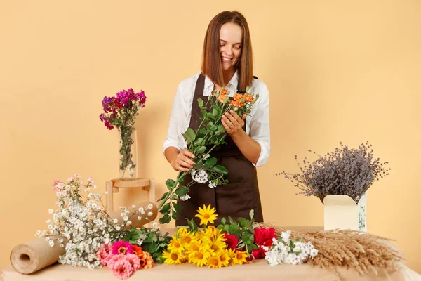 Flower bouquet. Gardener\'s service. Floral shop. Gift delivery. Beautiful blooms. Cheerful positive dark haired woman wearing brown apron posing isolated over beige background.