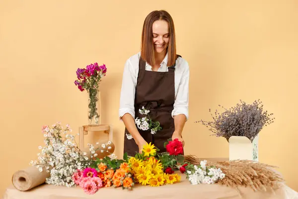 Florist occupation. Bunch delivery. Botanist job. Employee florist. Smiling dark haired woman wearing brown apron making bouquet posing isolated over beige background.