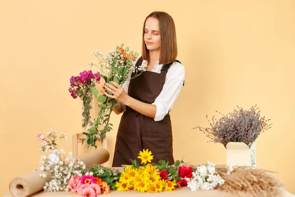 Gardening service. Florist worker. Floral store. People in the profession. Carrying flowers. Calm attractive dark haired woman wearing brown apron posing isolated over beige background.