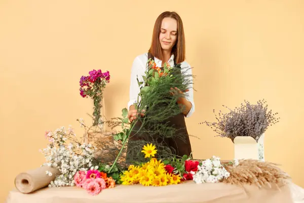 Working in a shop. Flower sale. Employee in gardening. Beautiful blooms. Making floral gifts. Charming calm smiling dark haired woman wearing brown apron posing isolated over beige background.