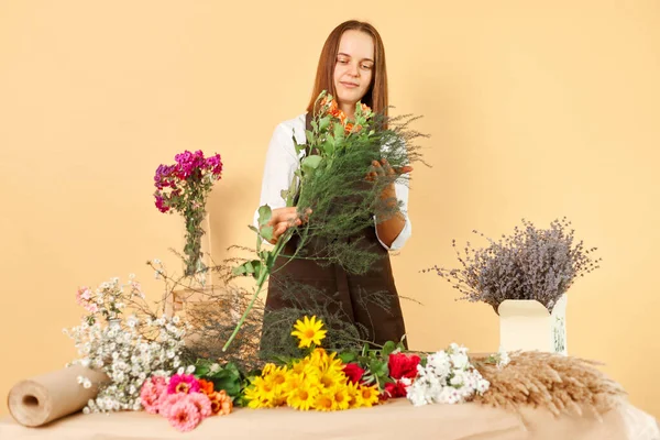 Florist\'s job. Bunch of flowers. Botanical delivery. Gardener\'s occupation. Floral arrangement. Positive dark haired woman wearing brown apron working with beauty posing isolated over beige background