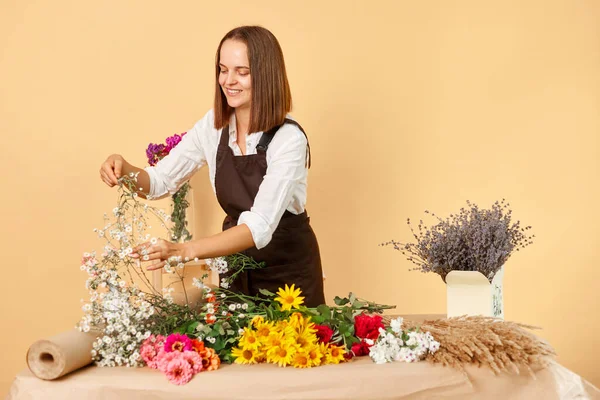 Working with flowers. Holding a bouquet. Sales profession. Flower advertising. Optimistic hardworking dark haired woman wearing brown apron posing isolated over beige background.