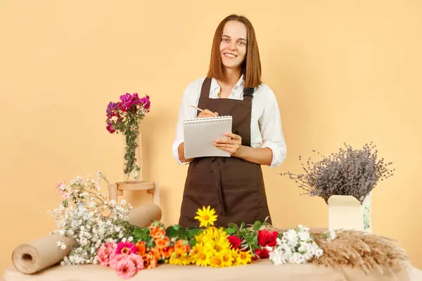 Bouquet decoration. Friendly florist. Business owner. Floral occupation. Gardener\'s job. Young woman flowers shop owner keeps records of orders wearing apron isolated over beige background.
