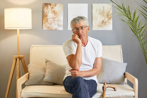 Bored sad upset grey-haired mature man wearing casual white T-shirt sitting on sofa at home interior felling boring holding chin thinking what to do looking at camera.