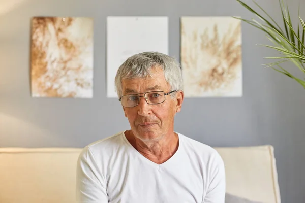 Calm grey-haired senior man in casual white T-shirt and glasses sitting on couch at home in living room interior looking at camera with focused expression.