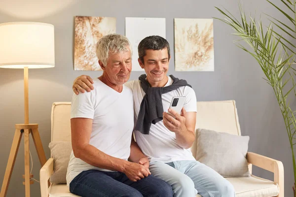 Happy senior gray-haired father and adult son using smart phone while sitting on sofa in living room family making selfie using mobile phone for online communication.