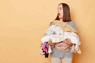 Çamaşırları tutan, bej arka planda izole edilmiş giysilerle dolu hayrete düşmüş bir kadın. Kirli giysi yığını reklamları için fotokopi odasına bakıyor..