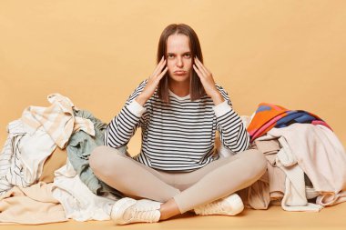 Bej arka planda soyutlanmış kıyafetlerle çevrili mutsuz bir kadın. Dolabını düzenlemek için yorgun, yorgun ve bitkin düşmüş baş ağrısı çekiyor..