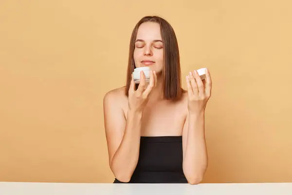 Satisfied young woman wearing black top holding container with moisturizer facial cream isolated over beige background smelling pleasant aromat of skin care product.