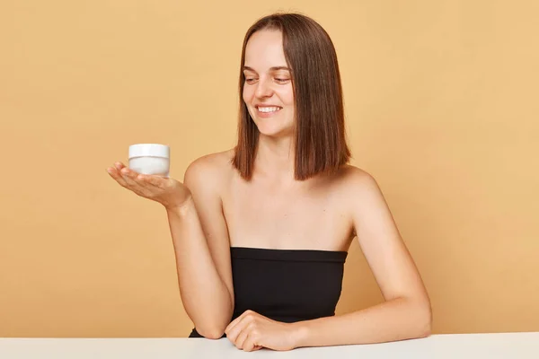 Face moisturizer. Skin care lifestyle. Salon treatment. Skin health and care. Charming happy young woman wearing black top holding container with facial cream isolated over beige background.