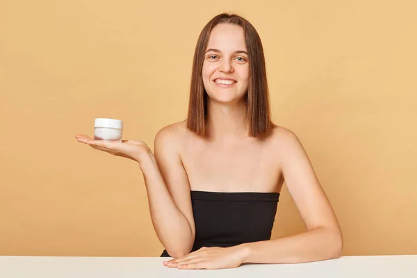 Health and beauty. Lifting skincare. Cute young woman wearing black top holding container with moisturizer facial cream on palm presenting new skin care products isolated over beige background.