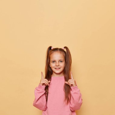 Pembe eşofmanlı neşeli küçük kız bej arkaplan üzerine izole edilmiş poz veriyor. Yukarıya ve boş alan düşman reklamına ya da tanıtım içeriğine işaret ediyor..