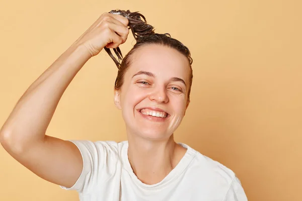Satisfied delighted woman washes hair applies shampoo taking shower isolated over beige background looking at camera with toothy smile doing beauty treatment for her hair.