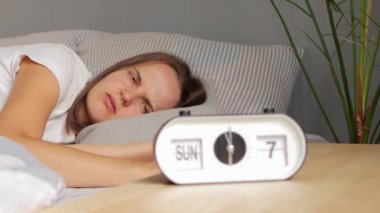 Kahverengi saçlı kızgın kadın saat 7 'de yatakta uzanıyor. Alarm çalıyor. Yüzünde negatif bir ifade var..