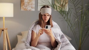Gözü bağlı mutsuz bir kadın elinde modern akıllı telefonuyla internette sohbet ediyor. Evde çizgili battaniyeye sarılmış yatakta kötü şok olmuş haber pozları okuyor..