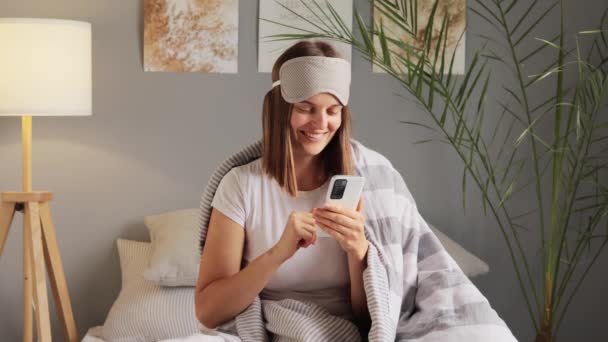 毛布に包まれた睡眠マスクを身に着けている幸せな女性は インターネットを介してオンラインビデオ通話を行います こんにちは またはさようなら 幸せを表現する彼女の友人に挨拶 フレンドリーな外観 — ストック動画