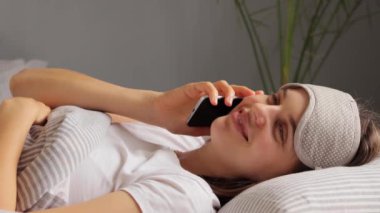 Gülümseyen neşeli kadın yatakta uzanıyor. Cep telefonuyla konuşuyor. Uyku maskesi ve beyaz tişört giyiyor..