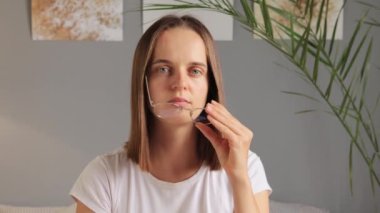 Kahverengi saçlı, yorgun bir kadın beyaz tişörtlü, evde oturuyor. Yorgun, yorgun, gözlüğünü çıkarıyor, gözlerini ovuşturuyor, acı çekiyor..