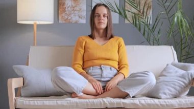 Rahatlamış bir kadın evinde, oturma odasında meditasyon yaparken bacak bacak bacak üstüne atmış poz verirken ve el ele dua ederken gözleri derin nefes alıp verişini ve konsantrasyonunu koruyor..